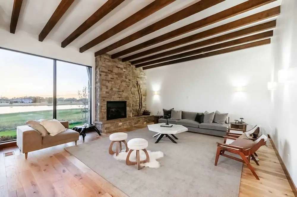 Living room with a stone fireplace, exposed ceiling beams, and large windows overlooking a lawn.